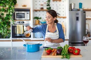 contento joven mujer Cocinando saboreo cena en un maceta en pie en moderno cocina a hogar. ama de casa preparando sano comida sonriente . casa y nutrición. dieta recetas concepto foto