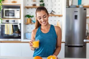 hermosa joven mujer Bebiendo Fresco naranja jugo en cocina. sano dieta. contento joven mujer con vaso de jugo y naranja a mesa en cocina. foto