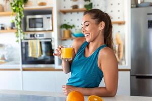 Beautiful young woman drinking fresh orange juice in kitchen. Healthy diet. Happy young woman with glass of juice and orange at table in kitchen. photo