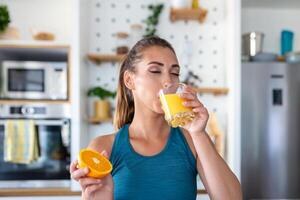 hermosa joven mujer Bebiendo Fresco naranja jugo en cocina. sano dieta. contento joven mujer con vaso de jugo y naranja a mesa en cocina. foto