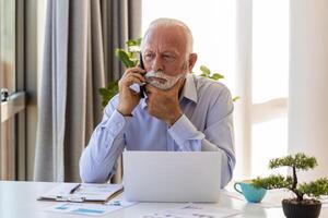 Mature businessman working on laptop and talking on cellphone. Handsome mature business leader sitting in a modern office photo