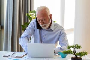 Mature businessman working on laptop and talking on cellphone. Handsome mature business leader sitting in a modern office photo