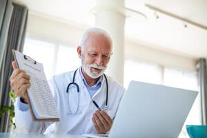 Serious old mature professional male doctor using laptop computer in hospital office having medical webinar training, writing in healthcare report, consulting patient online at telemedicine meeting. photo