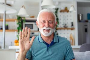 Smiling senior man wave to camera having video call on laptop, happy elderly male sit on couch at home talk using modern technologies and wireless connection photo