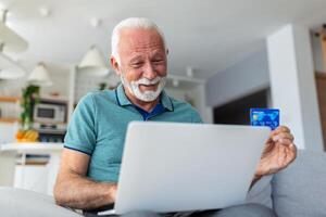 Mature man using laptop, holding plastic credit or debit card, senior grey haired customer making secure internet payment, shopping or browsing online banking service, entering information photo