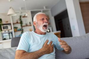 mayor hombre prensas mano a cofre tiene corazón ataque sufre desde inaguantable dolor, maduro hombre con dolor en corazón en vivo habitación. mayor hombre sufrimiento desde malo dolor en su cofre corazón ataque a hogar foto