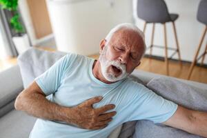 mayor hombre prensas mano a cofre tiene corazón ataque sufre desde inaguantable dolor, maduro hombre con dolor en corazón en vivo habitación. mayor hombre sufrimiento desde malo dolor en su cofre corazón ataque a hogar foto