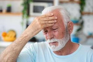 Tired, depressed senior man sitting on couch in living room feeling hurt and lonely. Aged, white-haired man touching forehead suffering from severe headache or recalling bad memories photo