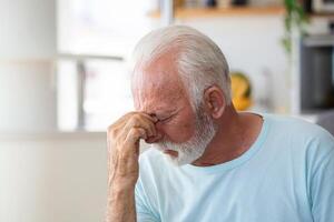 Tired, depressed senior man sitting on couch in living room feeling hurt and lonely. Aged, white-haired man touching forehead suffering from severe headache or recalling bad memories photo