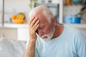 Tired, depressed senior man sitting on couch in living room feeling hurt and lonely. Aged, white-haired man touching forehead suffering from severe headache or recalling bad memories photo