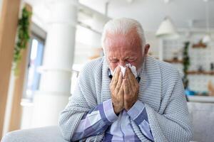 Senior sick bearded man sneezes into napkin at home on gray sofa with white blanket. Disease, protection, coronavirus, virus, disease, flu, respiratory dressing. photo