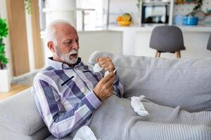 enfermo mayor hombre comprobación su temperatura sufrimiento desde estacional gripe o frío acostado en sofá riendo sufrimiento desde estacional gripe o frío. . enfermo mayor sensación insalubre con influenza a hogar foto