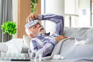 Mature man feeling sick with cold and fever at home, ill with flu disease sitting on the sofa with ice pack on his head photo