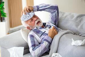 Mature man feeling sick with cold and fever at home, ill with flu disease sitting on the sofa with ice pack on his head photo