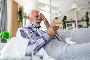 Sick elderly man checking his temperature suffering from seasonal flu or cold lying on sofa caughing suffering from seasonal flu or cold. . Ill senior feel unhealthy with influenza at home photo