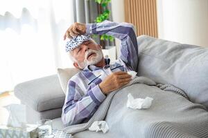 maduro hombre sensación enfermo con frío y fiebre a hogar, enfermo con gripe enfermedad sentado en el sofá con hielo paquete en su cabeza foto