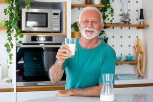 mayor hombre Bebiendo un vaso de Leche con un contento cara en pie y sonriente. hermoso mayor hombre Bebiendo un vaso de Fresco Leche en el cocina foto