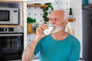 mayor hombre Bebiendo un vaso de Leche con un contento cara en pie y sonriente. hermoso mayor hombre Bebiendo un vaso de Fresco Leche en el cocina foto