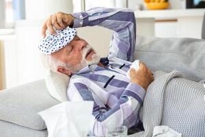 Mature man feeling sick with cold and fever at home, ill with flu disease sitting on the sofa with ice pack on his head photo