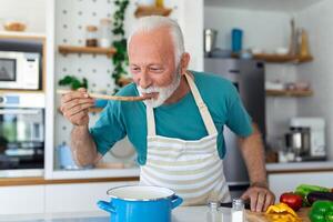 Happy retired senior man cooking in kitchen. Retirement, hobby people concept. Portrait of smiling senior man holding spoon to taste food photo