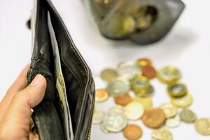 Hand holding black wallet with only one bank note inside and blurry coins open from the piggy bank laid out scattered on white background. This image signifies the use of money inexorably. photo