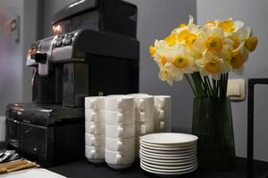 Many clean white cups and plates on the table near the coffee maker and vase with daffodils photo