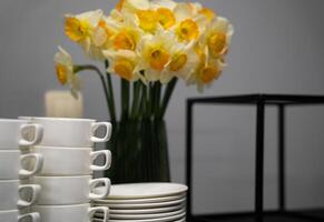 Many clean white cups and plates on the table near the coffee maker and vase with daffodils photo