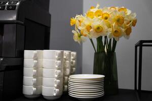 Many clean white cups and plates on the table near the coffee maker and vase with daffodils photo