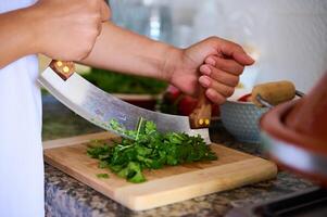 Close-up chef's hands using mezzaluna knife, chopping fresh green herb of parsley on a bamboo cutting board photo