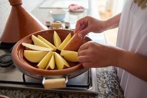 Moroccan tajine clay dish on the stove and Muslim woman housewife stacking slices of organic potato, cooking at home photo