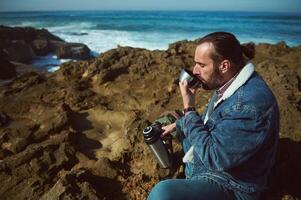 Bearded young man, tourist traveler adventurer sits on the rocky cliff and drinks water from thermos in nature by ocean photo