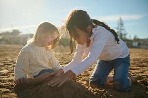 Two adorable little children girls playing together on the beach, building sandy castle on warm sunny day photo