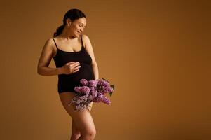 Smiling pregnant woman with a bouquet of lilacs, isolated over beige studio background, dressed in black lingerie photo