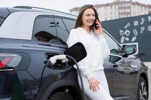 joven mujer cargando su eléctrico coche a un cargando estación en el ciudad. eco combustible concepto. el concepto de ambientalmente simpático transporte. recarga batería desde cargando estación. foto