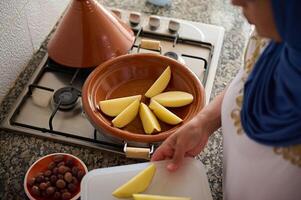 Moroccan tajine clay dish on the stove and blurred woman housewife stacking slices of organic potato, cooking at home photo