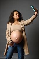 elegante embarazada mujer tomando selfie en teléfono inteligente, compartiendo su estilo de vida en último trimestre de el embarazo en social medios de comunicación foto