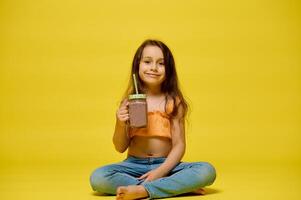 Smiling little girl drinks a cocktail from a glass bottle with a straw, isolated over yellow studio background. ad space photo