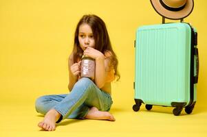 Beautiful little child girl drinking a cocktail, sitting near a trendy suitcase, isolated over yellow studio background photo