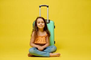 Caucasian cute traveler child girl with a suitcase, sitting over yellow studio background, smiling, looking at camera. photo