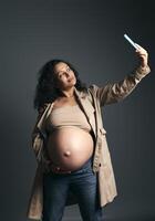 Happy pregnant woman takes self portrait on mobile phone while strokes her big belly, isolated fashion gray background photo