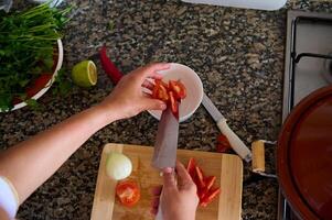 ver desde encima de un cocinero rebanar verduras, agregando Cortado Tomates dentro un blanco ensalada cuenco foto