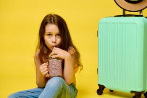 Adorable child girl drinking a fresh cocktail from straw, sitting near suitcase, isolated over yellow studio background. photo