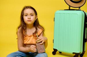 Lovely little kid girl drinking a cocktail, sitting near a trendy suitcase, looking at camera, isolated yellow backdrop photo