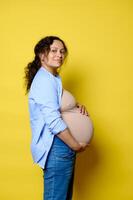 Happy pregnant woman putting hands on belly in last trimester of pregnancy, smiling at camera, isolated yellow backdrop photo