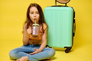 A kid girl drinking cocktail, sitting near a trendy suitcase, looking at camera, isolated over yellow studio background photo