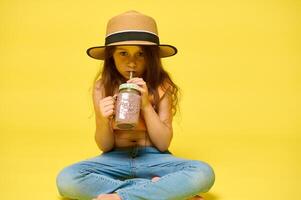 Cute stylish child girl drinks a cocktail from a glass bottle with a straw, isolated over yellow studio background photo