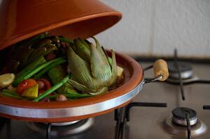 recortado ver de arcilla maceta tagine con abierto loid mientras vegetales son humeante en el cocina cocina. marroquí comida concepto foto