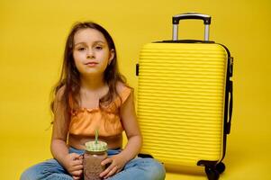 Cute child girl drinking a cocktail, sitting near yellow suitcase, looking at camera, isolated studio background photo