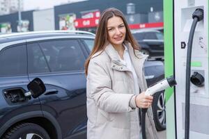 joven mujer cargando su eléctrico coche a un cargando estación en el ciudad. eco combustible concepto. el concepto de ambientalmente simpático transporte. recarga batería desde cargando estación. foto