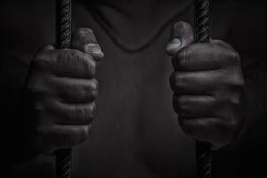 closeup on hands of man sitting in jail. Man behind jail bars on black background photo